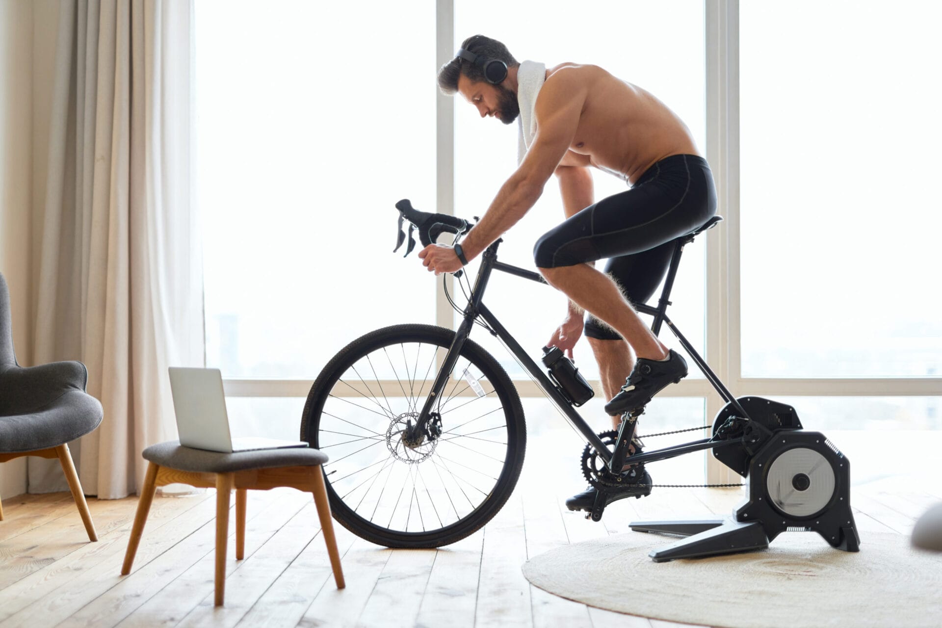 Handsome shirtless man in headphones enjoying favorite songs and grabbing bottle of water while riding exercise bicycle at home