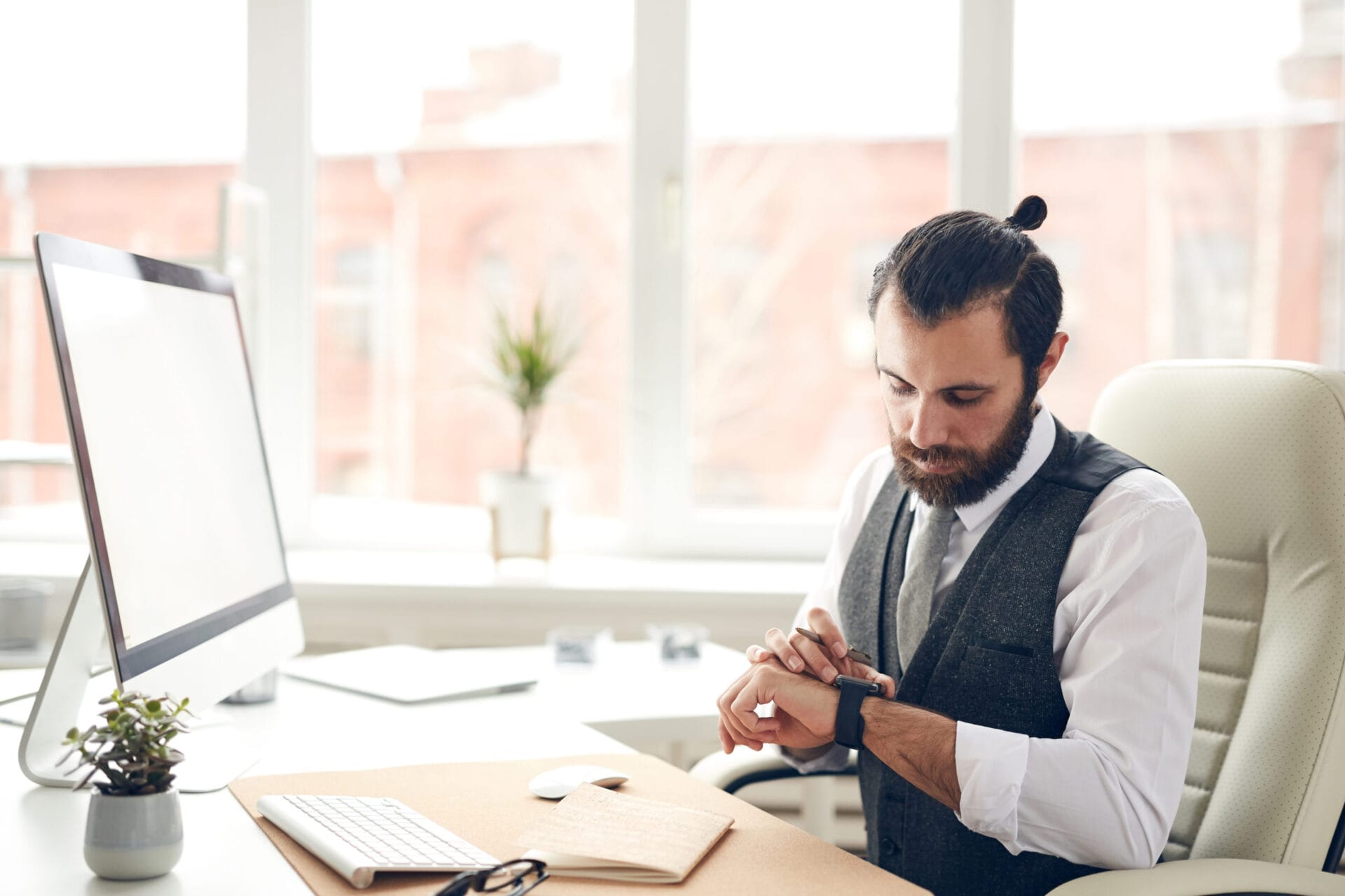 Serious handsome bearded businessman in waistcoat sitting at desk with computer and checking message on smart watch