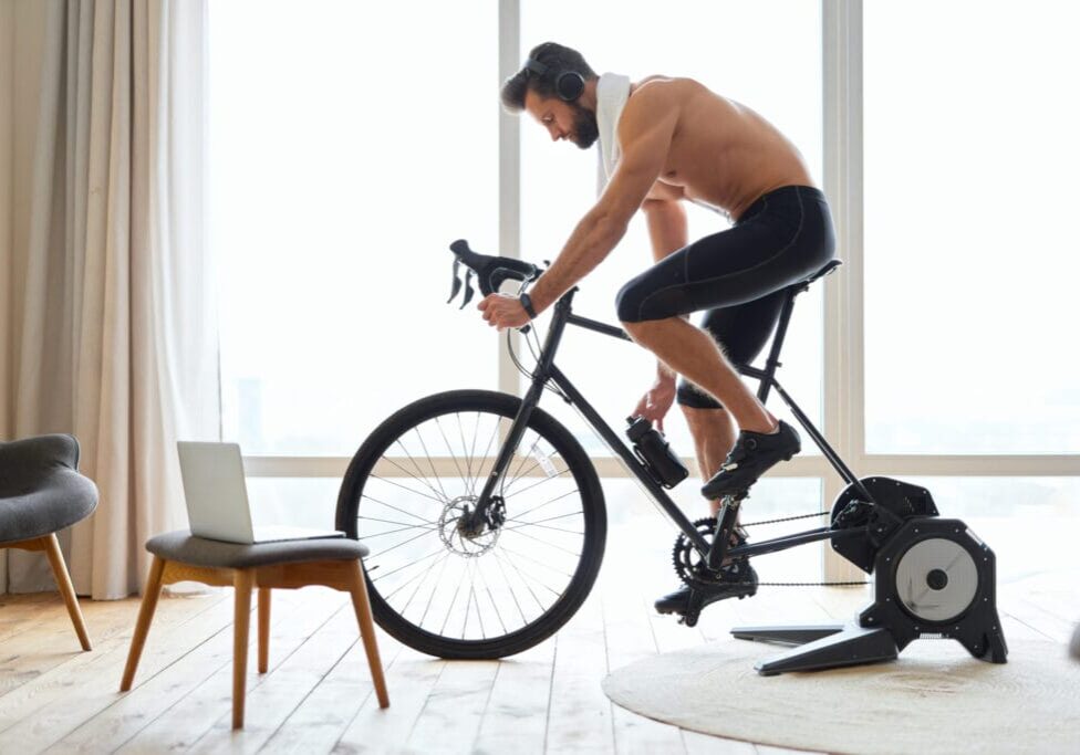 Handsome shirtless man in headphones enjoying favorite songs and grabbing bottle of water while riding exercise bicycle at home