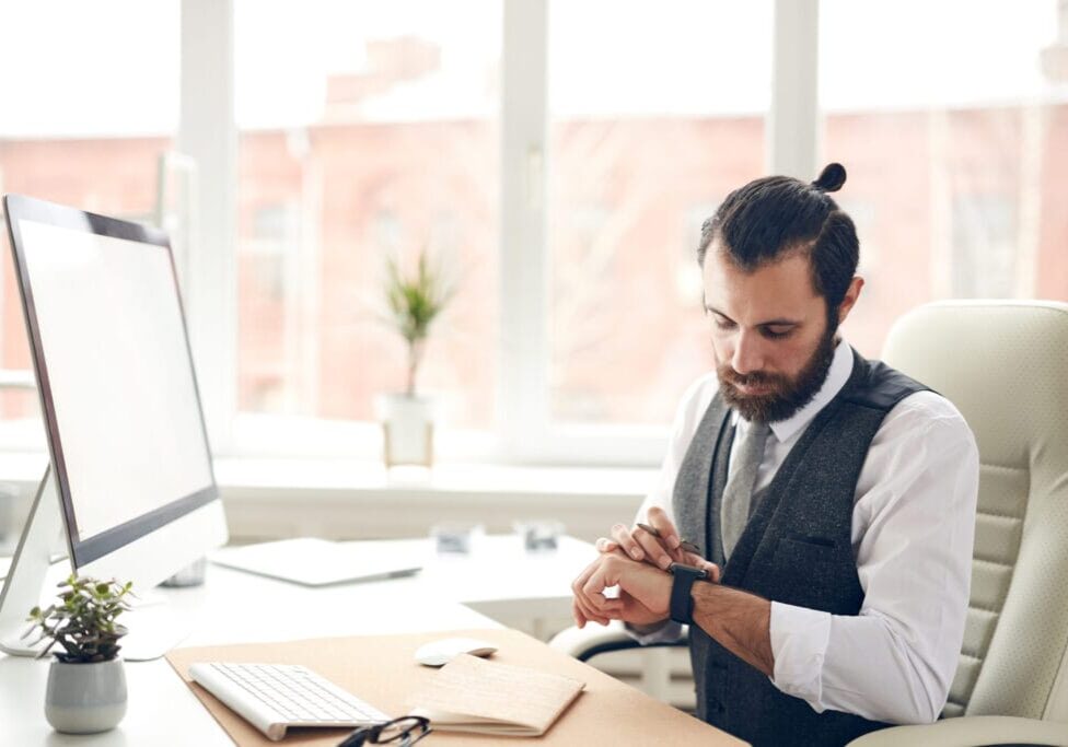 Serious handsome bearded businessman in waistcoat sitting at desk with computer and checking message on smart watch