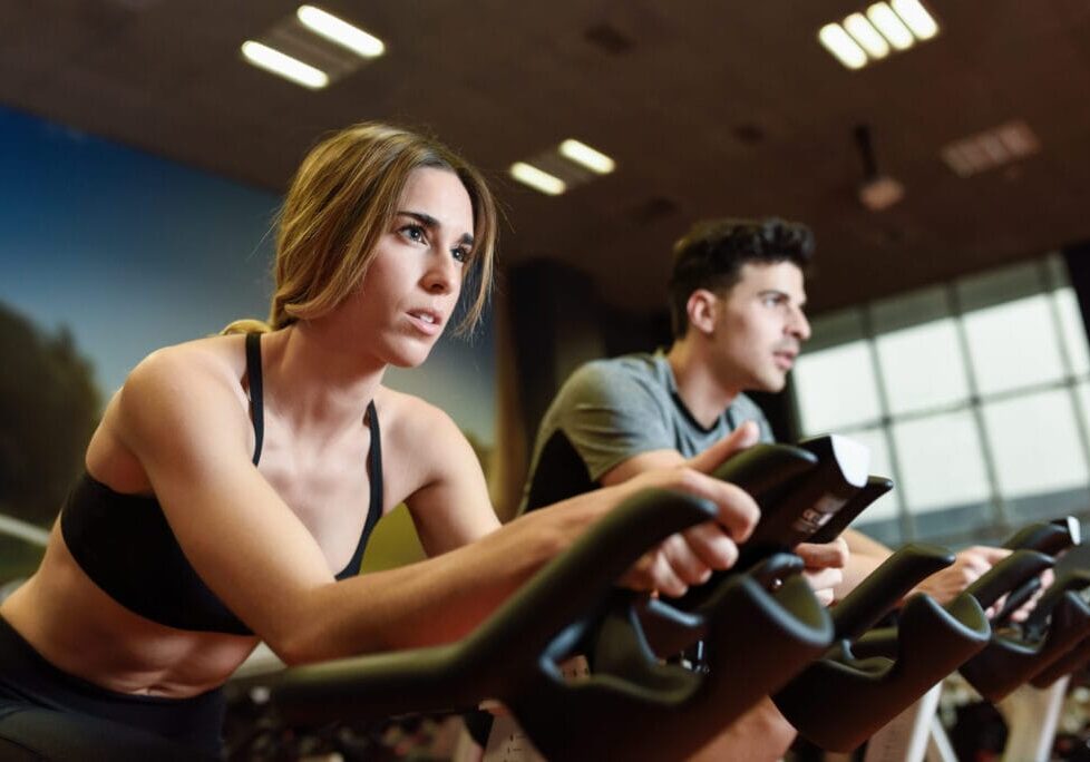 Attractive woman and man biking in the gym, exercising legs doing cardio workout cycling bikes. Couple in a spinning class wearing sportswear.