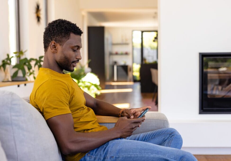 Happy african american man using smartphone in living room. Relaxation, lifestyle, communication and domestic life, unaltered.